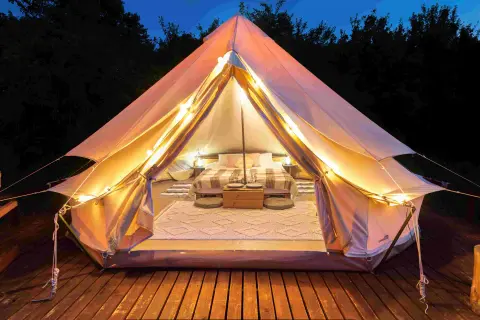 Przytulny namiot z łóżkiem i oświetleniem, otoczony drzewami. To jest właśnie glamping – luksusowy kemping.