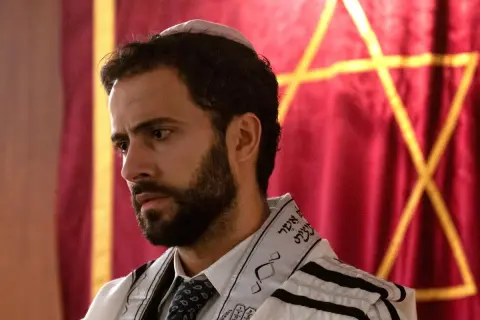 Ein Mann mit Bart und Kippa trägt einen Tallit. Im Hintergrund ist ein Davidstern auf rotem Stoff zu sehen. So sieht ein Jude aus.