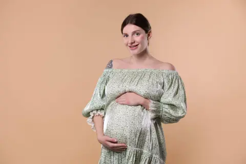 Une femme enceinte, au teint clair et aux cheveux bruns tirés en arrière, sourit à la caméra. Elle porte une robe verte à fleurs, dénudant ses épaules, et pose ses mains sur son ventre arrondi.