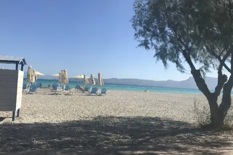 Słoneczny dzień na plaży Alkyon. Białe leżaki i parasole czekają na plażowiczów. W tle turkusowe morze i góry.