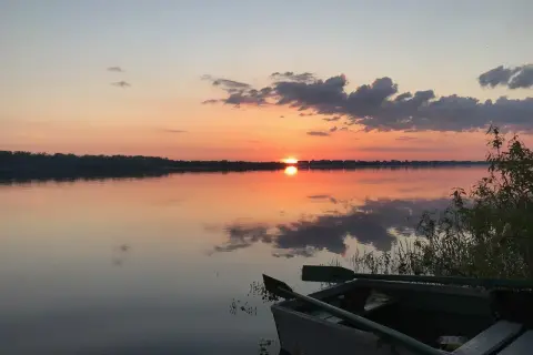 Łódź czeka nad spokojną rzeką białych nocy, odbijając pomarańczowe niebo i chmury.