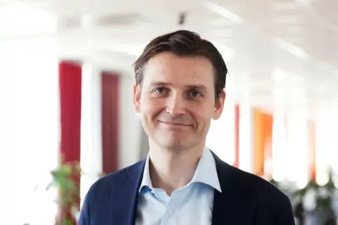 Peter Laurin, en man med blå ögon och mörkt hår, ler mot kameran i en mörkblå kavaj och ljusblå skjorta.