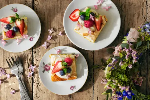 Tre tallrikar med vegansk midsommarmat, toppad med bär och blommor, ligger på ett träbord. En blomsterkrans ligger bredvid.