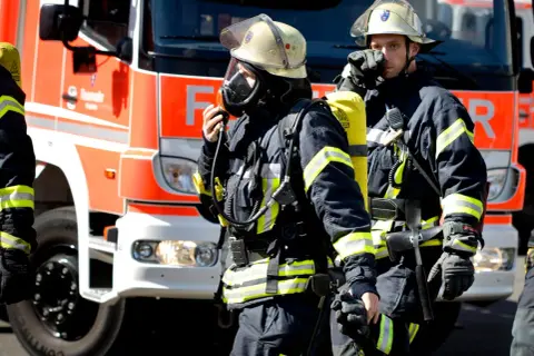 Zwei Feuerwehrmänner in voller Montur vor einem roten Feuerwehrauto. Sie sind bereit für den Einsatz. Wie viel verdient man als Feuerwehrmann?