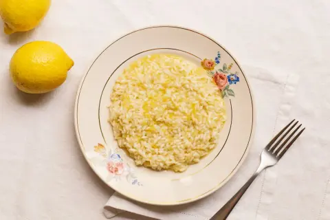 Un cremoso risotto al limon, con ralladura de limón espolvoreada, servido en un plato floral. Dos limones frescos reposan cerca.