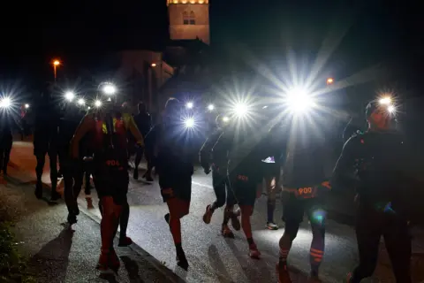 Courseurs de trail de nuit, lampes frontales allumées, traversant un village sombre. Un chien les accompagne.