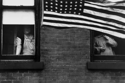 Une photographe femme américaine capture deux femmes regardant par des fenêtres, un drapeau américain flottant au-dessus.