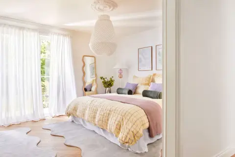 Ein geschwungener Spiegel im Schlafzimmer reflektiert das helle Zimmer mit Bett, weißen Vorhängen und Blick ins Grüne.