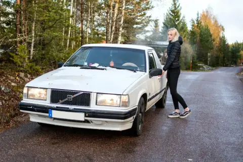 Vit Volvo med en tjej som öppnar dörren. En svensk registreringsskylt syns tydligt på bilen.