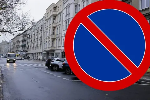 Znak B35 zakazu zatrzymywania się na mokrej ulicy z samochodami i kamienicami w tle.