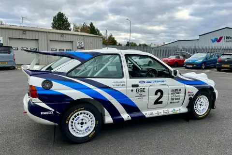 En vit rallybil Ford Escort Cosworth med blåa detaljer och nummer 2. Bilen står parkerad utomhus.