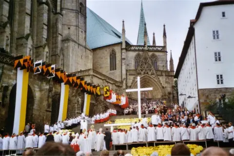 Große Menschenmenge vor einer Kathedrale, die an einem Hochamt teilnimmt. Fahnen und Girlanden schmücken die Fassade.