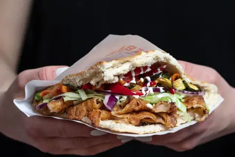 Kebab w bułce z mięsem, warzywami i sosami, trzymany w dłoniach.