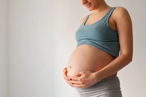 Uśmiechnięta ciężarna kobieta trzyma dłonie na brzuchu. Czas na poród, może już niedługo pojawi się czop śluzowy.