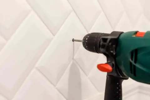 Taladro perforando un agujero en una pared con azulejos blancos para colocar tacos.