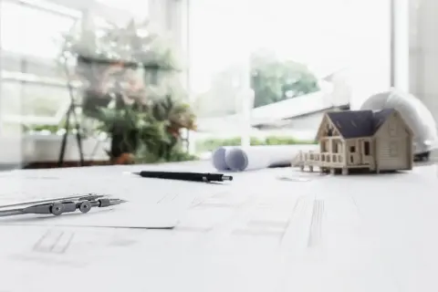 Model domu, cyrkiel i plany architektoniczne na stole. W tle okno z zielenią. Projekt uwzględnia nieprzekraczalna linia zabudowy.