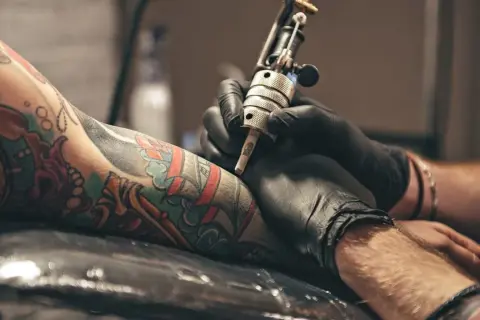 Manos enguantadas trabajan en un tatuaje colorido en un brazo. El artista usa una máquina de tatuar, creando arte permanente.