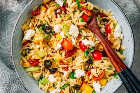 Ein bunter Kritharaki Salat mit Tomaten, Oliven, Feta und Paprika, serviert in einer Schüssel mit Holzstäbchen.