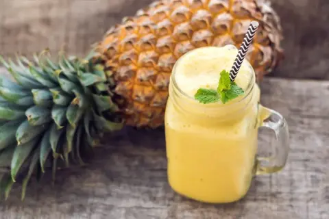 Orzeźwiający smoothie z ananasem, ozdobione miętą, obok świeżego owocu na drewnianym tle.