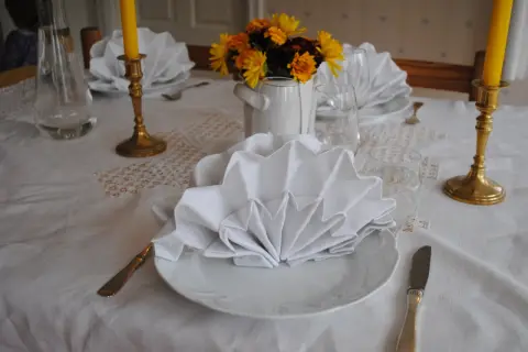 Vackert dukat bord med vita, vikta servetter i glas. Guldljusstakar och blommor skapar en festlig stämning.