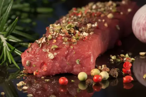 Un pezzo di carne succulenta, condita con pepe misto e rosmarino, pronta per diventare una deliziosa carne tipica pugliese. Accanto, uno spicchio d'aglio.