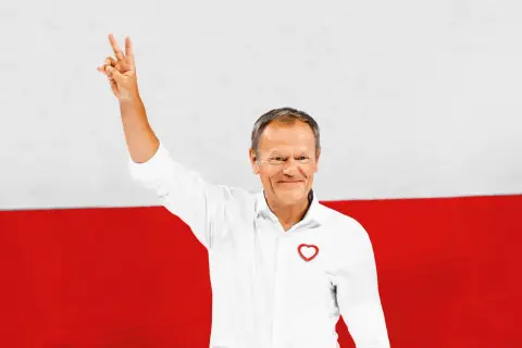 Donald Tusk z podniesioną ręką, symbolizującą zwycięstwo, na tle flagi Polski. Jego biała koszula z sercem i uśmiech podkreślają pozytywny przekaz. Polityka zagraniczna Koalicji Obywatelskiej w jego wizji.