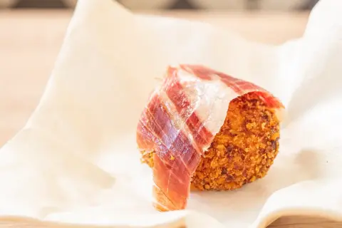 Croquetas de jamón crujientes, envueltas en una loncha de jamón serrano, servidas sobre papel blanco.