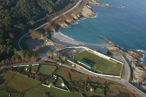 Vista aérea de un campo de fútbol junto a una playa a marosa, con el mar azul y rocas.