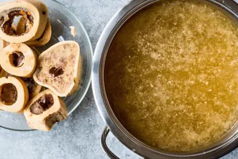 Un plato con huesos de tuétano y una olla con caldo dorado, perfecto para preparar caldo de huesos para perros.