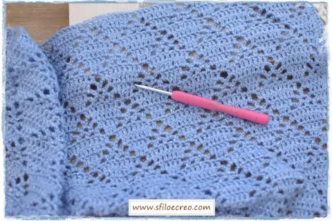 Un uncinetto rosa riposa su un lavoro a maglia azzurro con fili scintillanti, mostrando un delicato punto incrociato uncinetto.