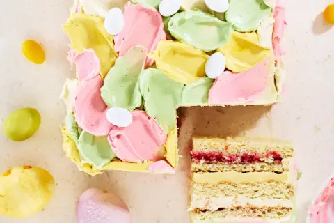 En vacker vit choklad tårta med pastellfärgad frosting och påskägg. En bit är uppskuren och visar lager av kräm och sylt.
