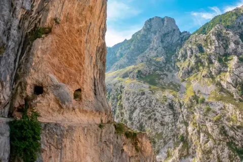 Senderos tallados en roca, con personas caminando. El inicio y fin de la ruta del Cares se pierden entre imponentes montañas verdes y cielos azules.