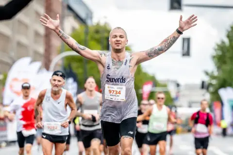 Triumfujący biegacz z tatuażami, z rękami w górze, na maratonie poniżej 2h.