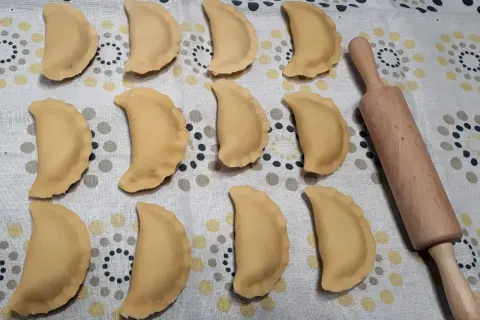 Gotowe do gotowania pierogi z jabłkami, ułożone rzędami na wzorzystym obrusie, obok drewnianego wałka.