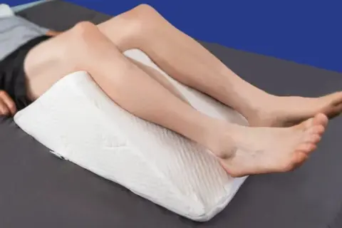 A person is resting with a white wedge pillow under their knees, providing comfort for sleeping with pillow under knees.
