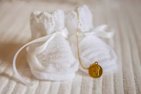 Douces chaussons blancs tricotés, ornés de rubans, avec une médaille dorée de la Vierge. Un symbole de marraine sans baptême, plein d'amour.