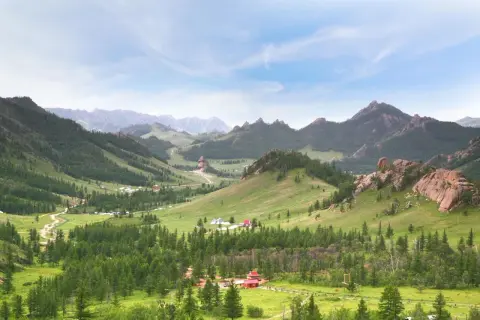 Grüne Hügel, Nadelwälder und Felsformationen prägen diese weite mongolische Landschaft. Kleine Gehöfte und Jurten sind in der Ferne zu erkennen.