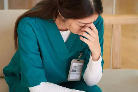 Une infirmière en blouse verte, le visage caché dans ses mains, semble lutter pour trouver son équilibre émotionnel.