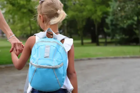Dziecko w niebieskim plecaku trzyma za rękę dorosłego, idąc przez park.