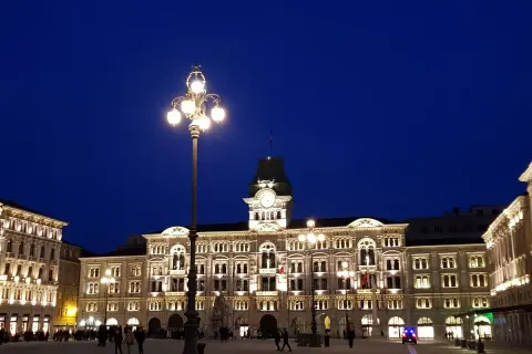 Piazza Unità d'Italia a Trieste, illuminata di sera. Edifici storici e lampioni creano un'atmosfera magica.