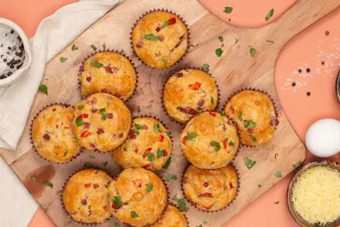 Herzhafte Pizza Muffins mit Speck und Paprika, garniert mit Petersilie. Daneben Zutaten wie Eier, Käse und Gewürze.