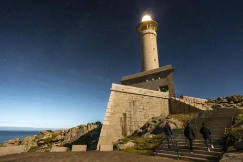 El faro de Punta Nariga ilumina la noche estrellada. Tres personas suben las escaleras hacia la torre, bajo un cielo lleno de estrellas.