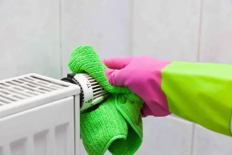 Mano con guanto rosa e verde pulisce il termostato del termosifone con un panno verde. Guida su come pulire i termosifoni.