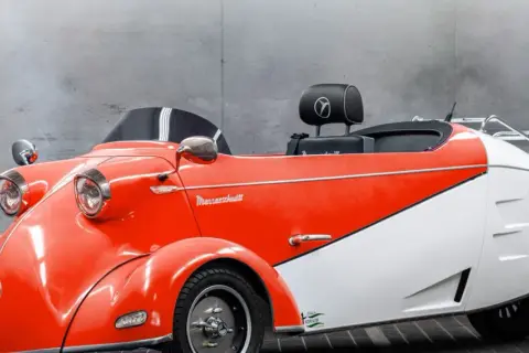 Ein leuchtend rotes und weißes Messerschmitt Auto mit Retro-Charme steht vor einer Betonwand.