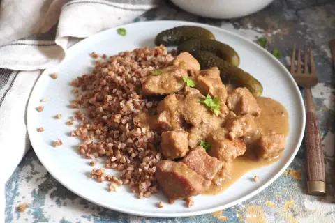 Gulasz z szynki wieprzowej w sosie własnym, podany z kaszą gryczaną i ogórkami kiszonymi. Pyszne, domowe danie.