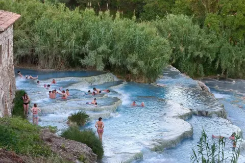 Persone si rilassano nelle cascate naturali dei aspio terme, con vegetazione lussureggiante e un antico edificio in pietra.