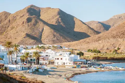 Vista de la finca Maltes del Fraile, con casas blancas, palmeras y montañas áridas al fondo. Barcas en la playa y el mar azul.