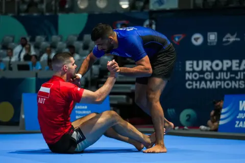 Två män tävlar i ju jitsu. En sitter på mattan i röd dräkt, den andra står i blå dräkt och sträcker ut en hand.
