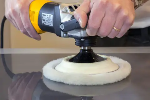 Manos puliendo una superficie oscura con una pulidora eléctrica. El disco de lana blanco gira para pulir madera.