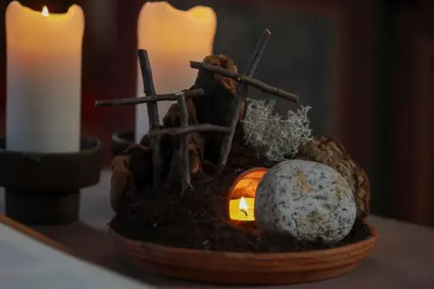Grab und Kerzenlicht symbolisieren die **heilige Woche**. Ein Stein liegt vor dem Eingang einer Höhle, aus der ein Licht scheint.